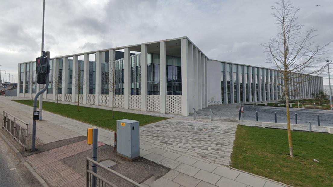 The court is box shaped and light grey in colour. It has large glass windows behind tall concrete pillars.
