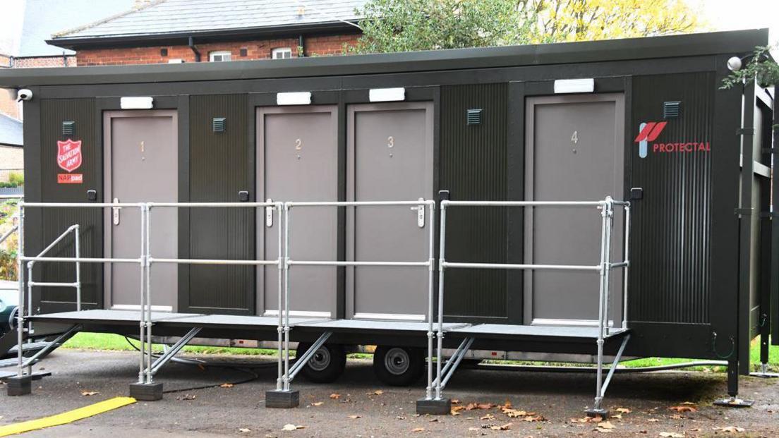 The shipping container-sized temporary building is black and grey with "The Salvation Army" branding on it. There are four doors which are numbered.