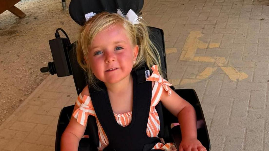 A young girl with blonde hair wearing a striped orange and white playsuit. She is sat in a black wheelchair with black straps holding her in. She is smiling and look at the camera.