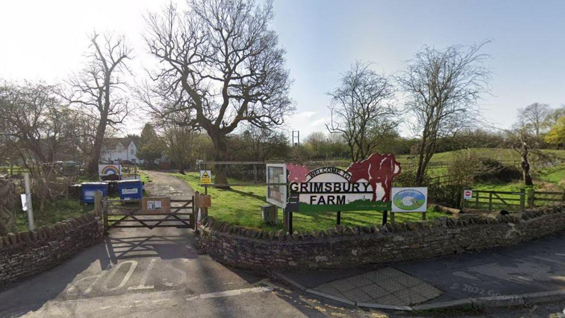 A photograph of a wooden farm gate with a low stone wall either side. A large sign indicates it is the entrance to Grimsbury Farm in Bristol. A driveway leads to a distant farm house. There are green fields and a mixture of trees and hedges behind the wall.