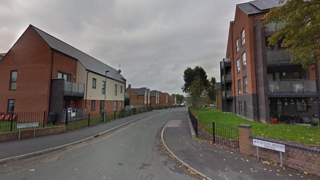 A junction with signposts reading Burton Wood Drive with a two-storey building on the left and a four-storey building on the right of a road