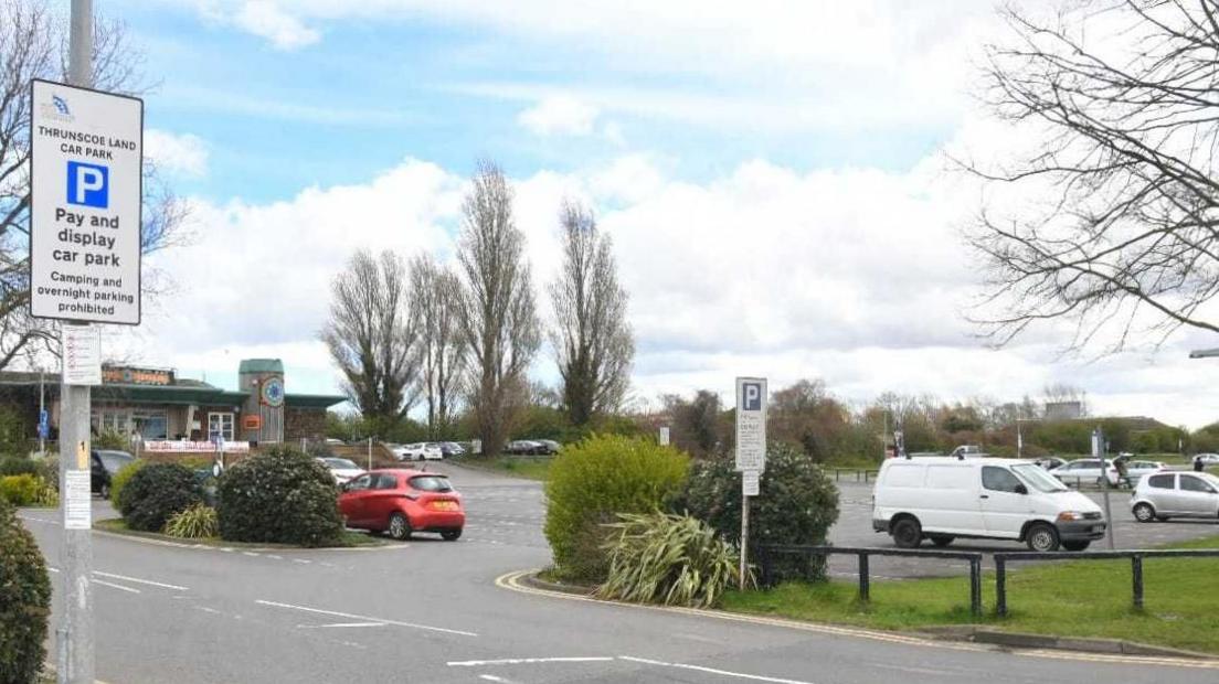 Pay and display sign at the entrance to the car park, with vehicles parked in bays and a building and trees in the background