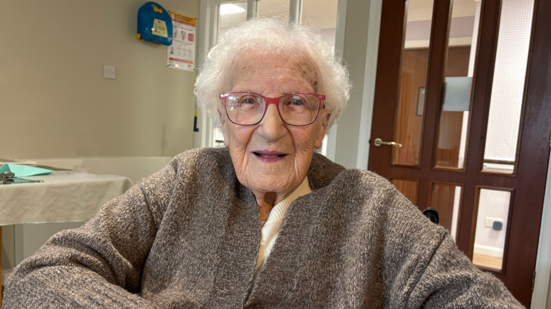 An elderly woman, 100-year-old May O'Shea, smiles into the camera. She has short, white curly hair, blue eyes, red glasses and is wearing a brown cardigan.