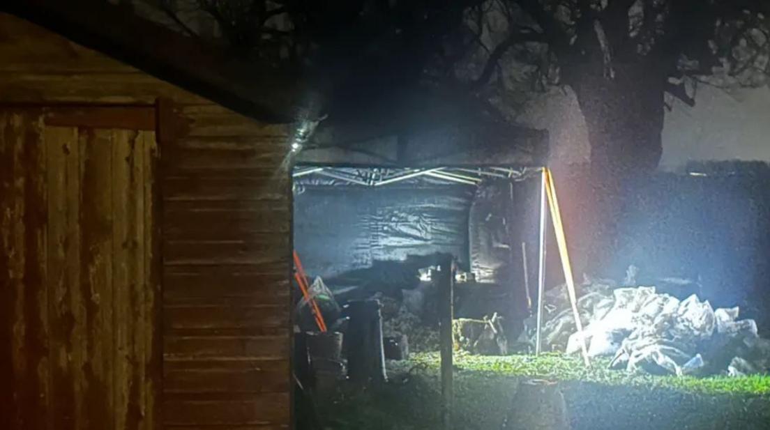 A forensic tent stands on a grassed area behind a wooden shed. A number of bags are visible on the right of the image. The sky is dark.
