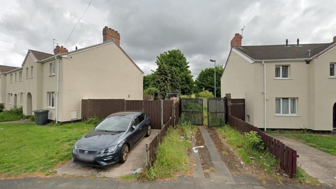 A pathway leads between two houses to a gate, partially open and which appears to lead to an area with greenery. A car is parked on the driveway of a nearby house.