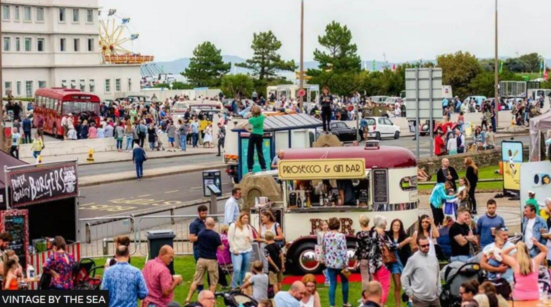 Crowds of people attend the festival, with a burger stall and a prosecco and gin bar. A vintage red bus is parked in the background.