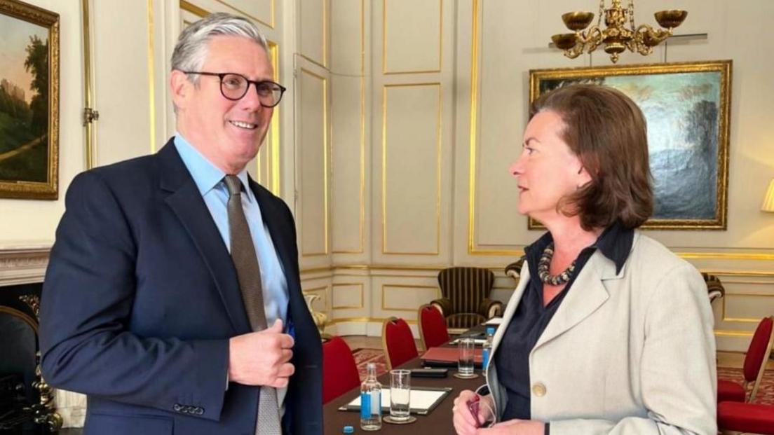 Sir Keir Starmer and Eluned Morgan stood in a meeting room with a chandelier, red chairs, a table and paintings hung on a wall.