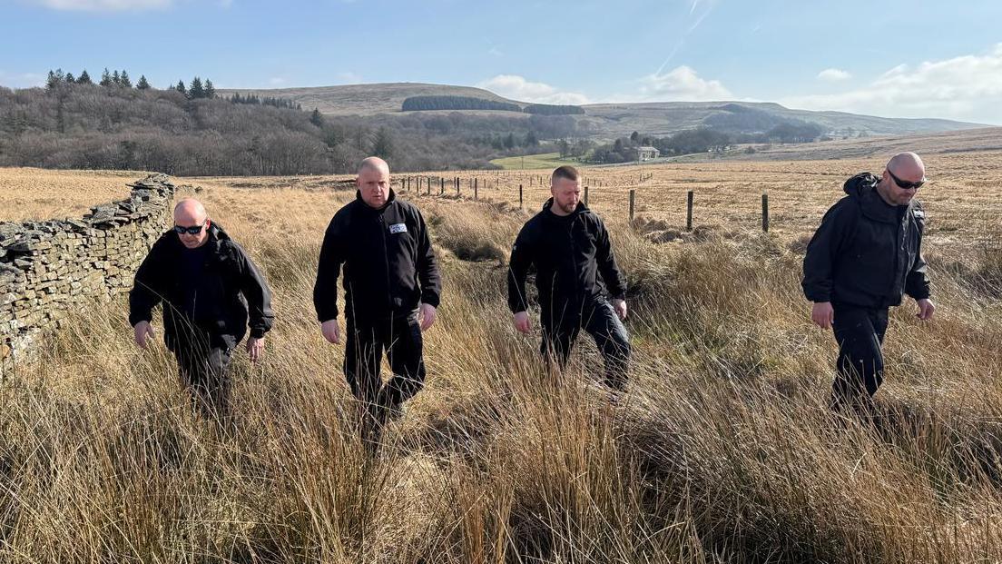 Four men dressed all in black walk through long, dry grass in between a wire fence and stone wall through a sloped field. Moorland and forest can be seen in the distance on a bright, sunny day. 