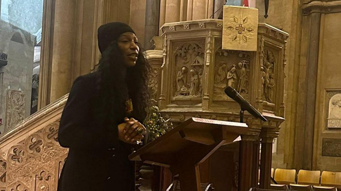 A woman called Jen Reid stands at a lectern with a microphone as she addresses an audience at Bristol Cathedral. She is wearing an all-black outfit including a black woolly hat