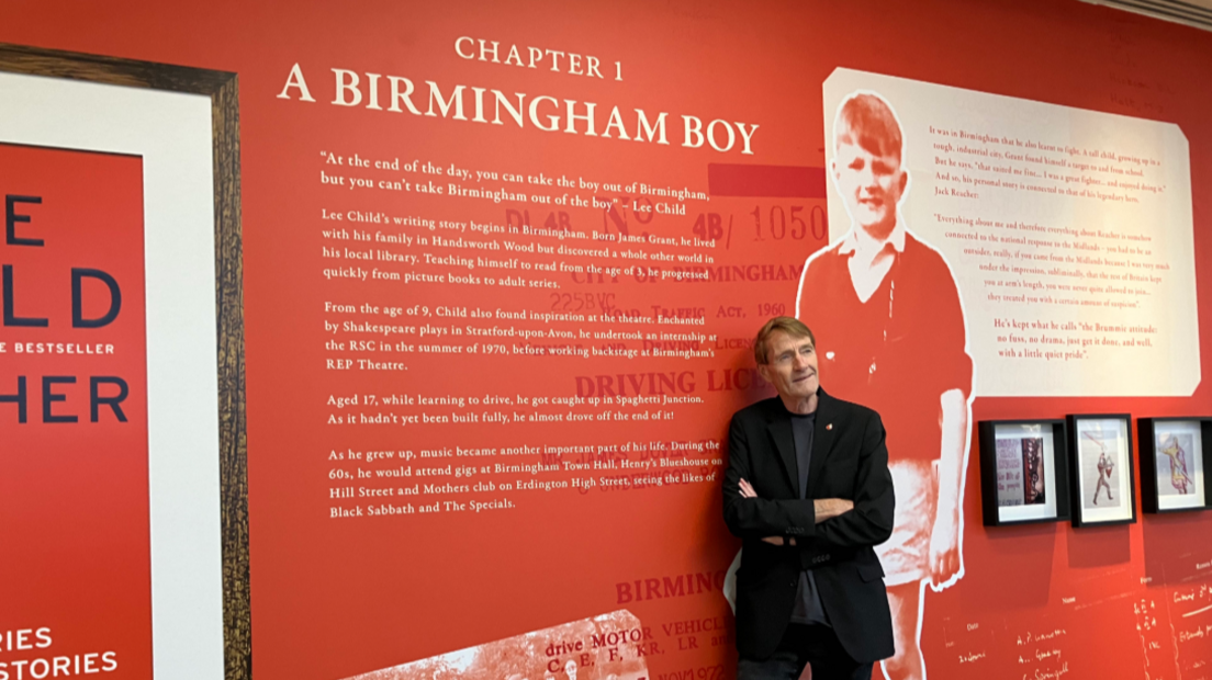 A man in a black suit type jacket, his arms crossed, leans against a display showing a large image of himself as a young boy. There is writing on the display, entitled 'Chapter 1 A Birmingham Boy'.