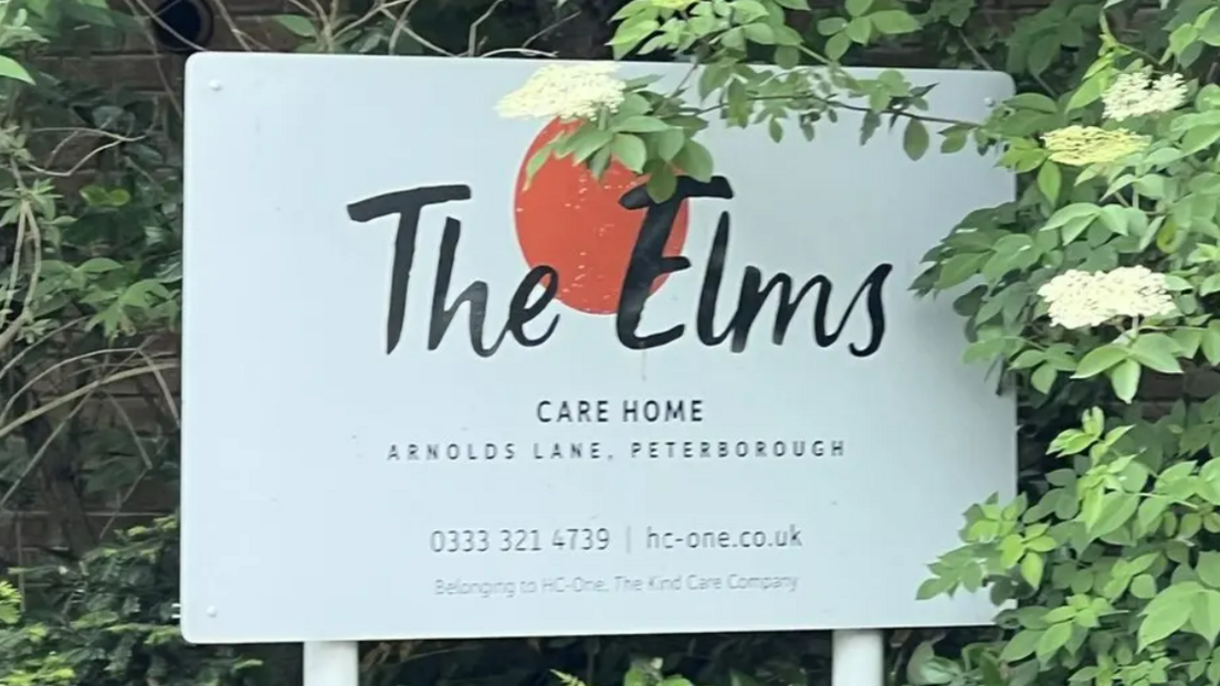A white sign in front of a bush which reads 'The Elms care home - Arnolds Lane, Peterborough'.