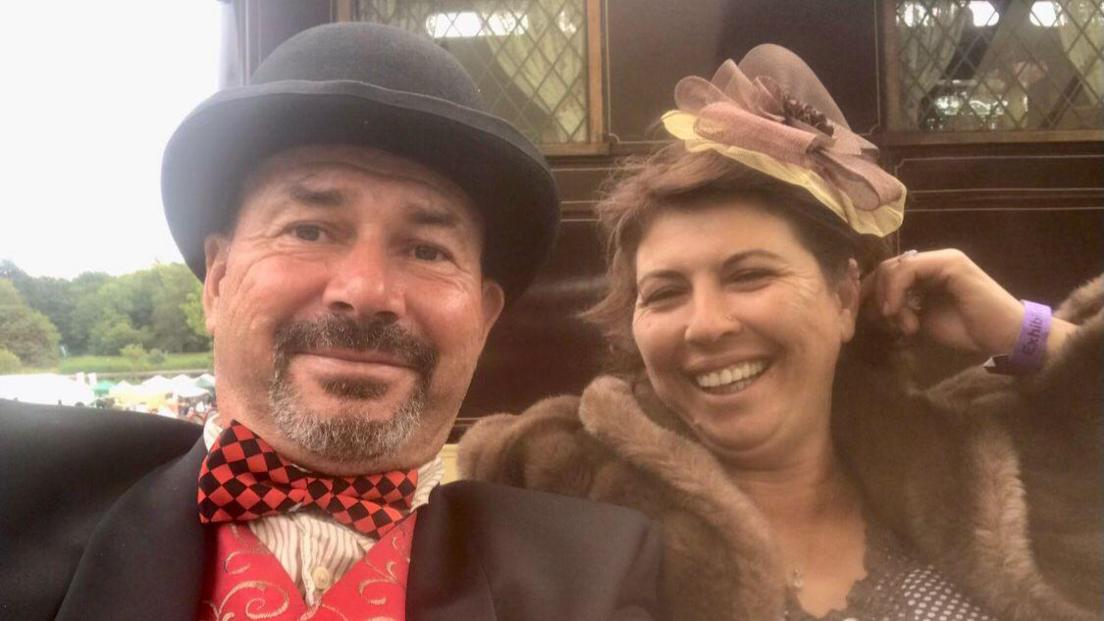 A man in a suit wearing a hat and natty bow tie sits next to a woman who is laughing in a big fur coat. 