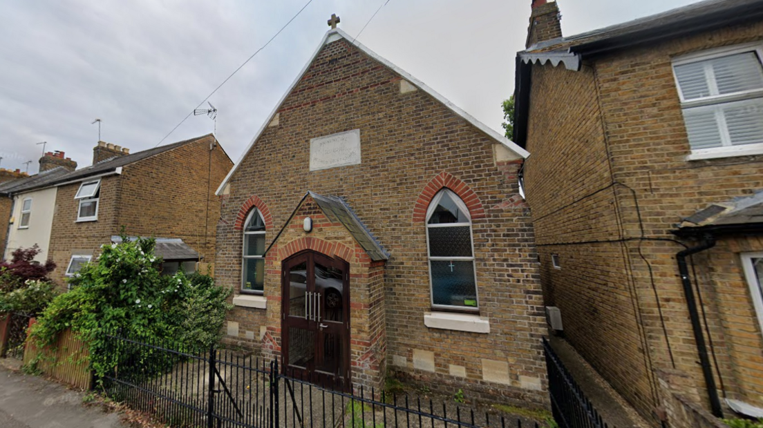 A Methodist church building. There is a small cross on the roof. The doors are brown wood and it has two lancet windows. The building sits in between terraced residential houses.