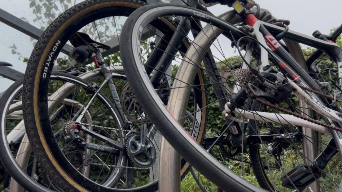 Close-up view of several bicycles parked together under a shelter. The image focuses on the wheels, gears, and frames, showing details like tires with brand markings, metal spokes, and chain mechanisms. Green foliage is visible in the background.