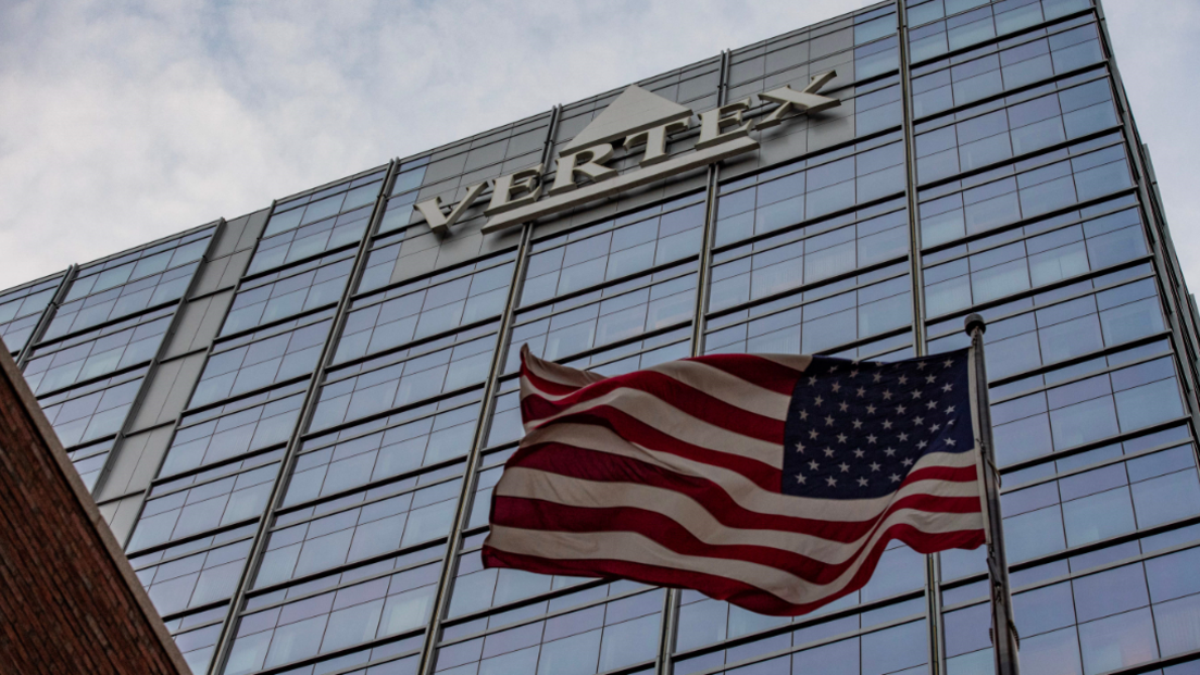 A stock image of the headquarters of Vertex Pharmaceuticals in America. The USA flag flutters in the wind in the forefront of the image.