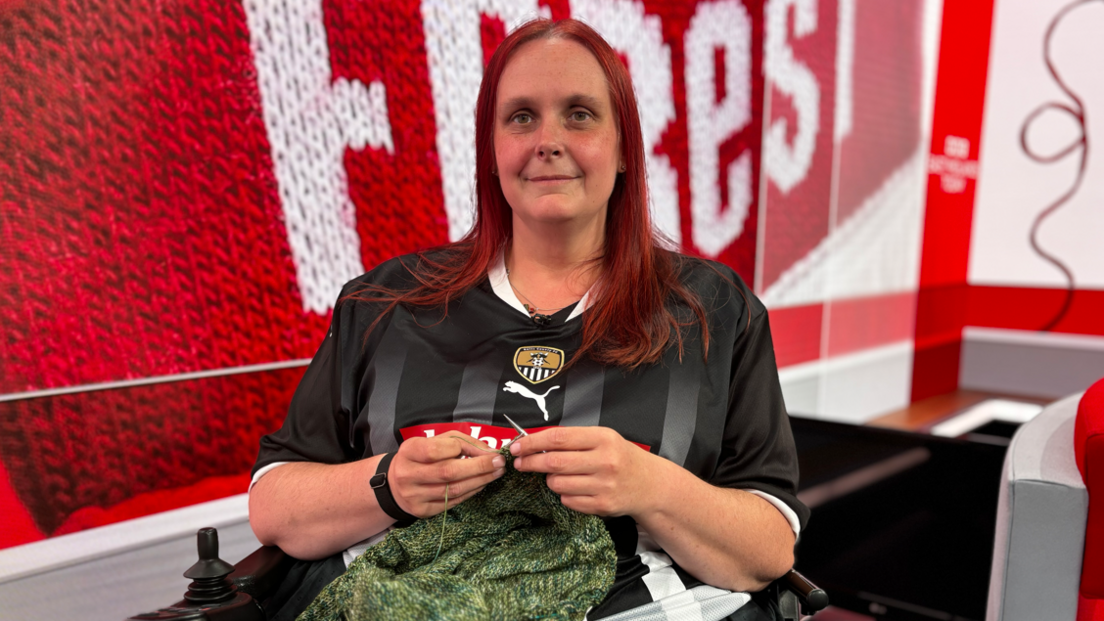 Ms Cameron knitting while wearing a Notts County shirt, sat in front of a Nottingham Forest-themed backdrop