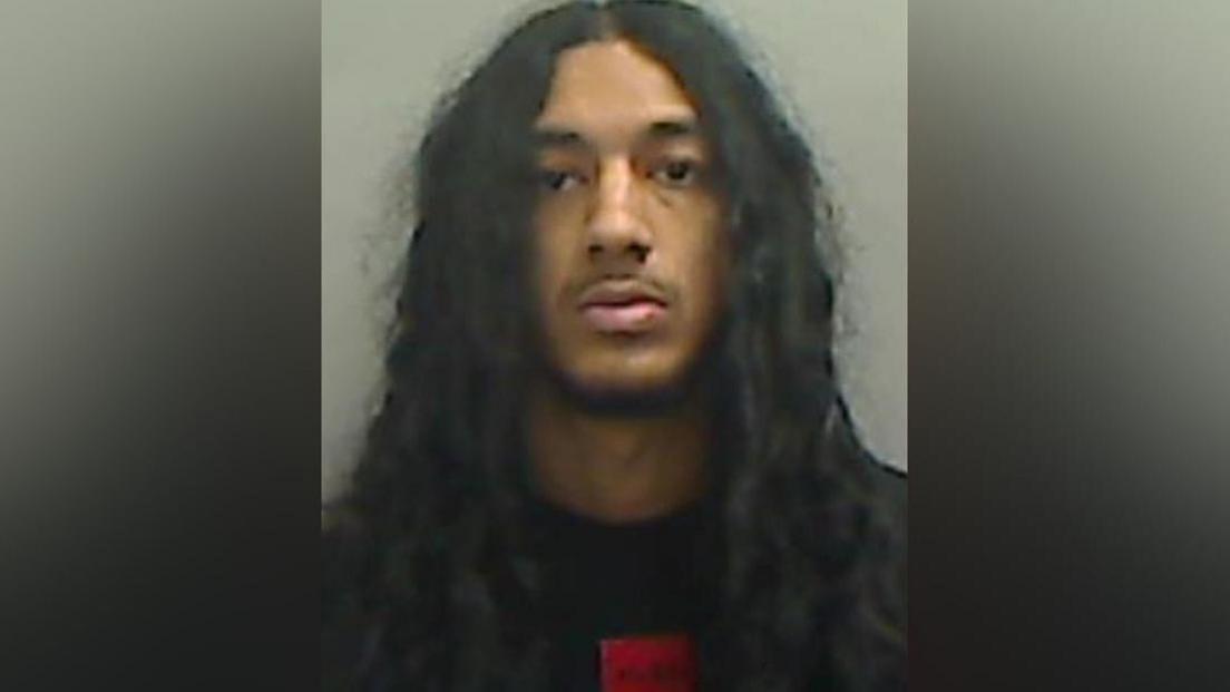 Mugshot of Mirza. He has very long thick black hair, a thin black beard and is wearing a black jumper with a red square logo beneath the neckline.