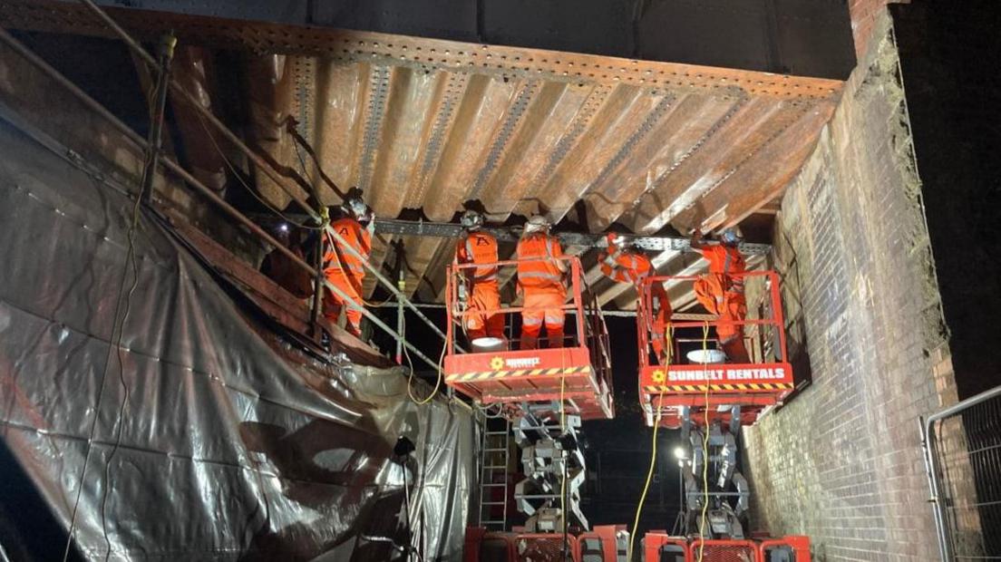 Workers in orange high-vis suits are working on the underside of a railway bridge. It is dark and only lit up by artificial lights.