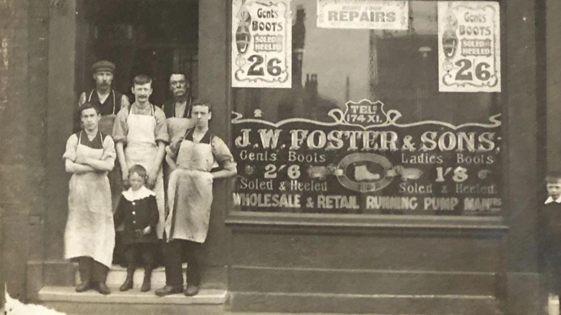 Five men, who are all wear aprons, and a young child are standing outside a shop in this black and white photograph. There are various advertisements for shoes in the window and along with the name of the firm: J.W. Foster and Sons. 
