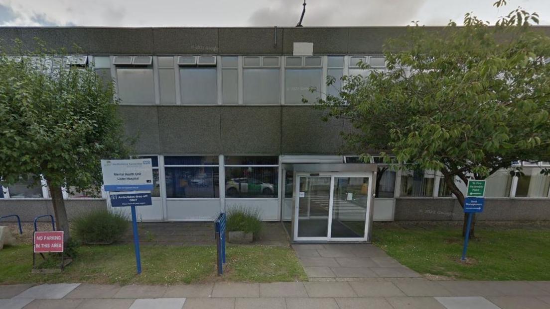 The exterior of the pre-fabricated two-storey mental health unit in the grounds of Lister Hospital in Stevenage. A sign reads 'Mental Health Unit, Lister Hospital'. There is an entrance with two sets of glass automatic doors.