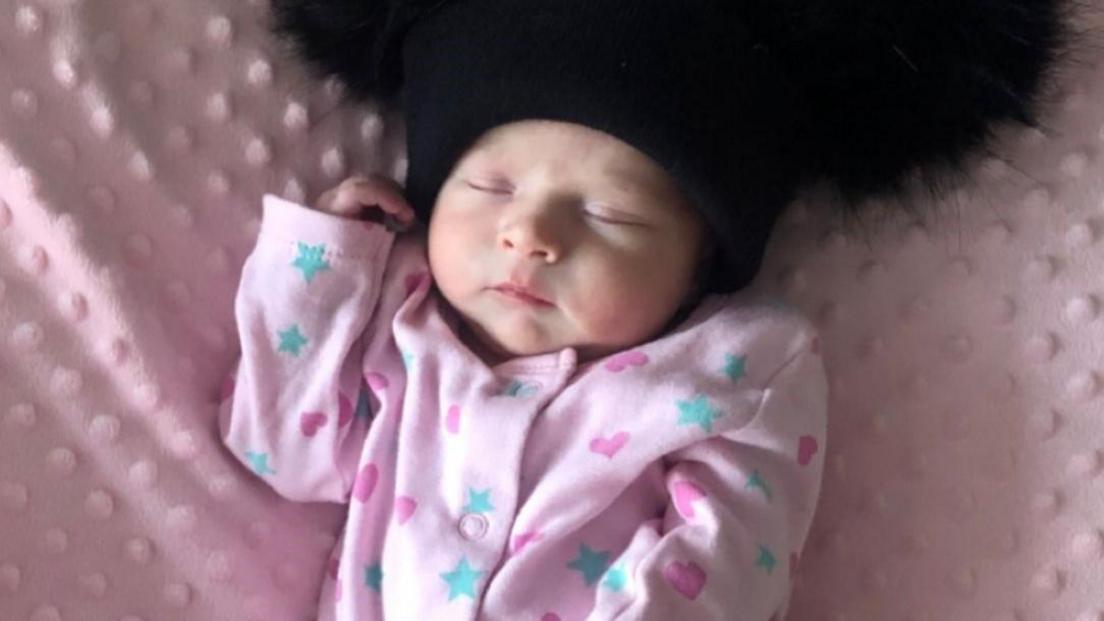 A baby wearing a pink babygrow with blue stars and dark pint hears on it. She is asleep while lying on a pink blanket and wearing a black hat