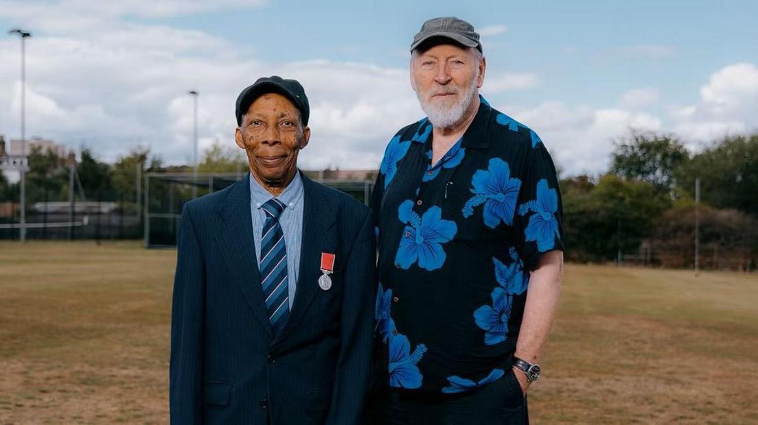 Siggy Cragwell, Thameslink's oldest employee, on the left, wearing a dark blue blazer and cap, a lighter blue shirt and a navy and blue striped tie. There is a British Empire Medal attached to his blazer. He is standing with Richard Thompson at a cricket ground. Mr Thompson has a white beard and is wearing a grey cap and black short-sleeved shirt with large blue flowers on.