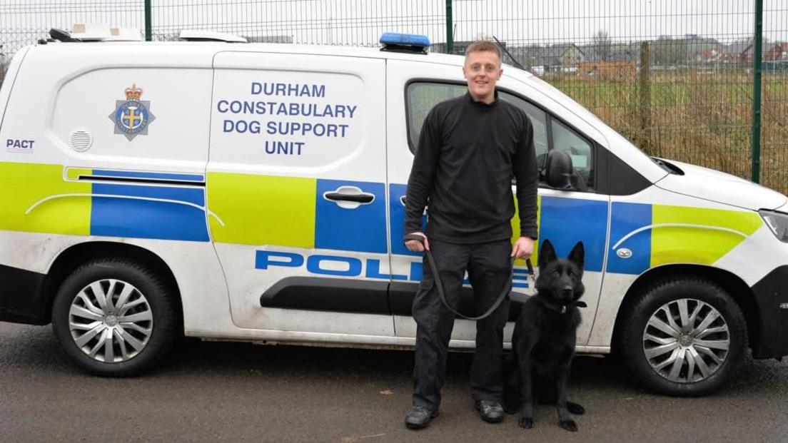 A black police dog with his handler stood in front of a police van