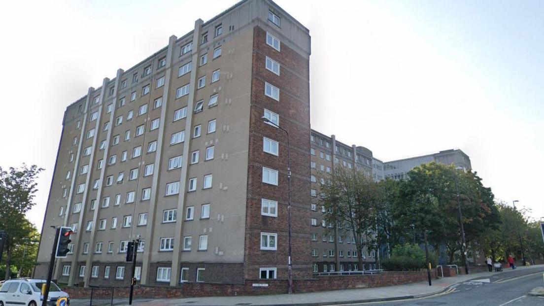 Streetview of a 10-storey block of flats. It is a combination of red brick and concrete. On the narrower side there are single windows on each floor. On the longer side there are 10 windows per floor. The block sits on a corner next to a set of traffic lights. 