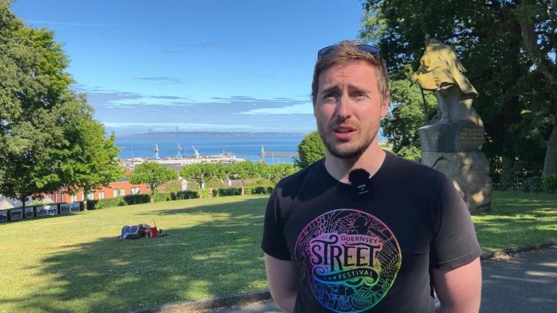 A man in a black t-shirt stands in a park, with the sea and other Channel Islands behind him