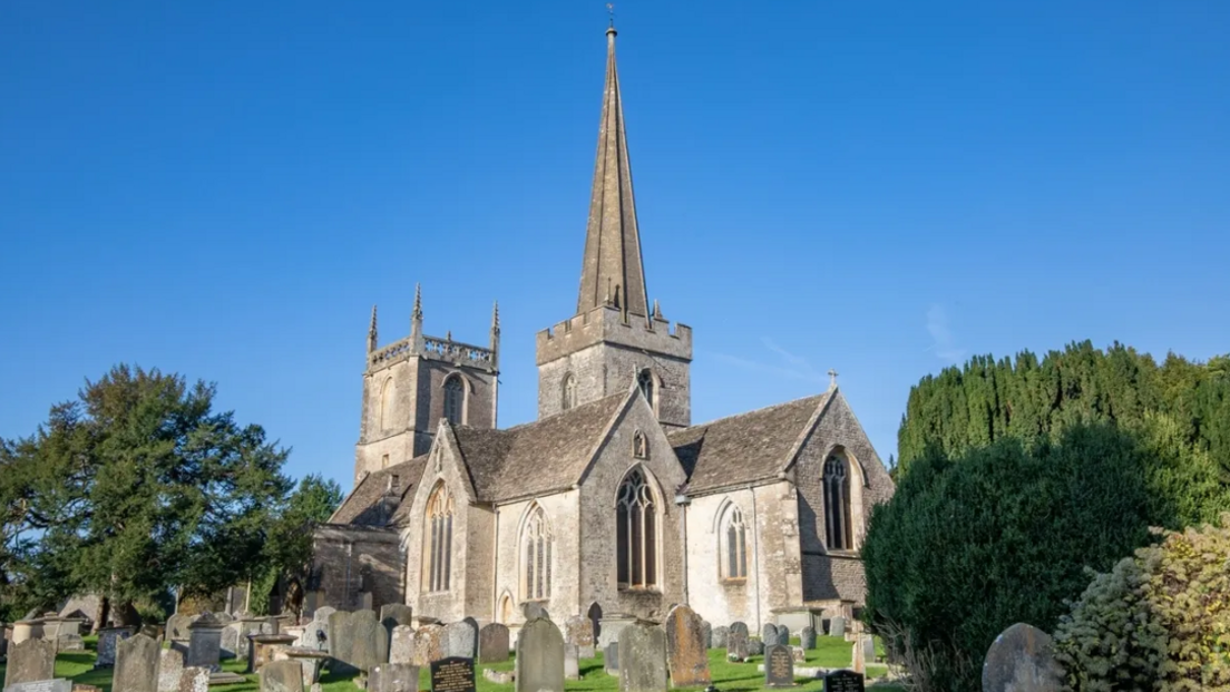 A medieval church on a sunny day. The church has a square tower and a spire. Around the church are many gravestones. 