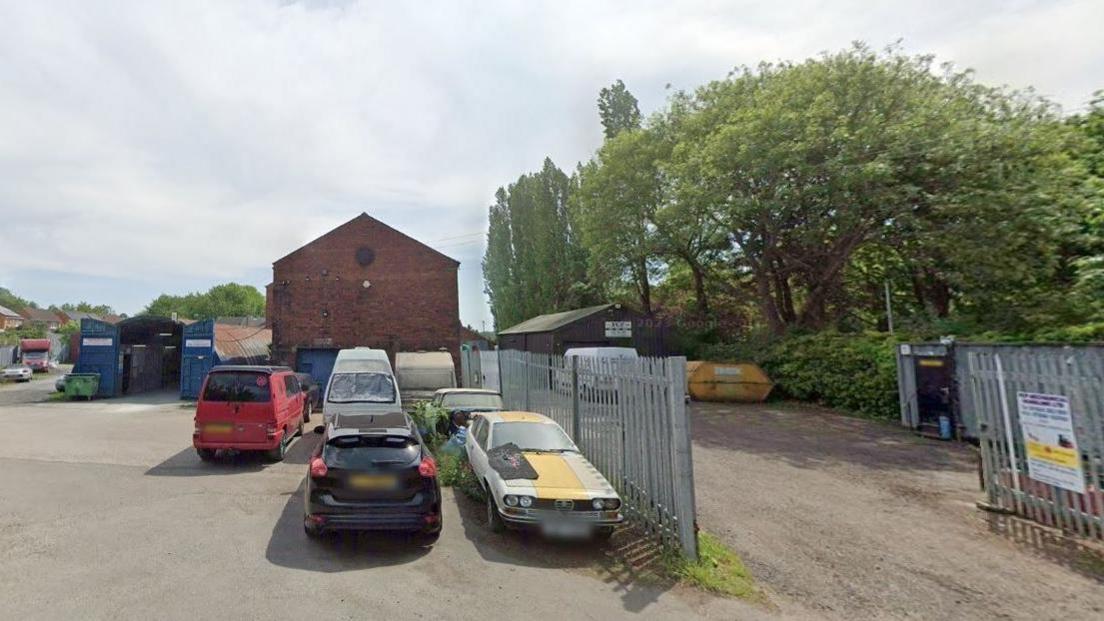 A car park with industrial buildings in the background and four cars 