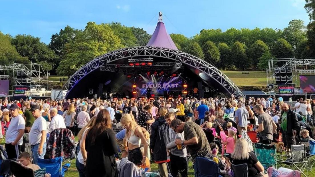 A large crowd gathered in front of a stage in Lower Castle Park. The stage is semi-circle shaped and has a purple roof. There are hundreds, if not thousands, of people stood in front of it.