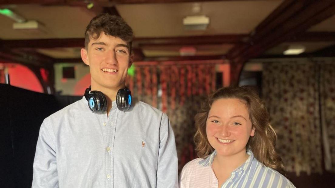 A young boy in his early twenties next to a young women also in her early twenties. The boy is very tall, with brown hair and blue eyes, he is wearing a blue shirt and has big black headphones around his neck. The girl is wearing a stripy blue and pink shirt, she has curly brown hair and is smiling at the camera.