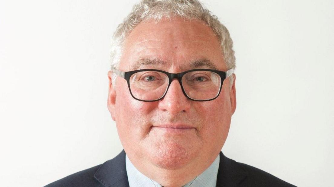 The photo shows Councillor Alan Preest. He is a white man with white hair, which is cropped short. He wears black rimmed glasses, and has a slight smile. He wears a dark suit jacket, with a light checked shirt.