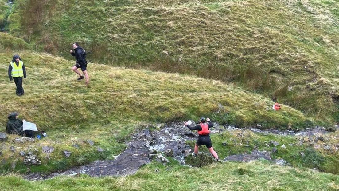 A runner is approaching a stream with a marshall in a high vis jacket watching on, above is another runner climbing the hill.