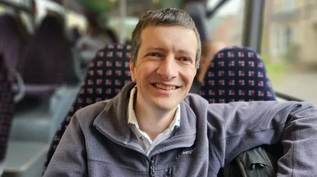 Picture of Andrew Cowell on a bus. He is wearing a grey fleece jacket. 