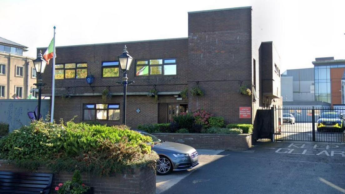 A brick building with a lantern outside showing the gardai logo. An Irish tricolour flag is next to a lamppost. A garda car is in the background.