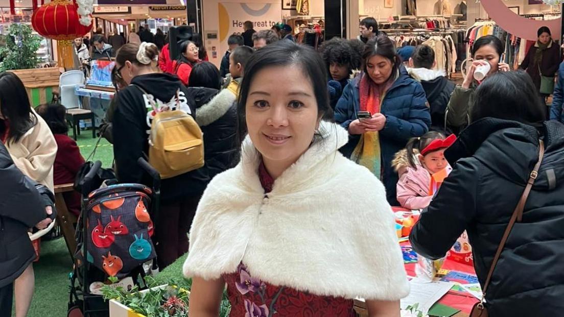 A woman with tied back dark hair smiles at the camera. She is wearing a white fur stole and a red floral dress. She is standing in a shopping centre.