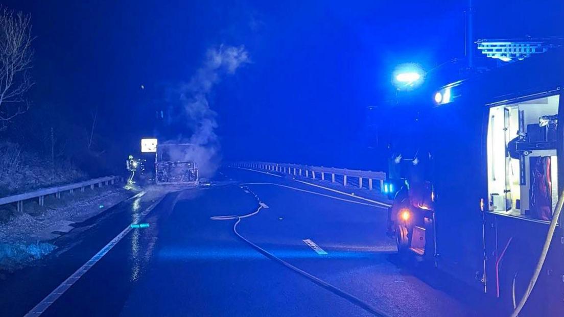 Smoke bellowing from a bus on the left with a firefighter standing beside it. There is a fire engine on the right with a hose running from it down the road.