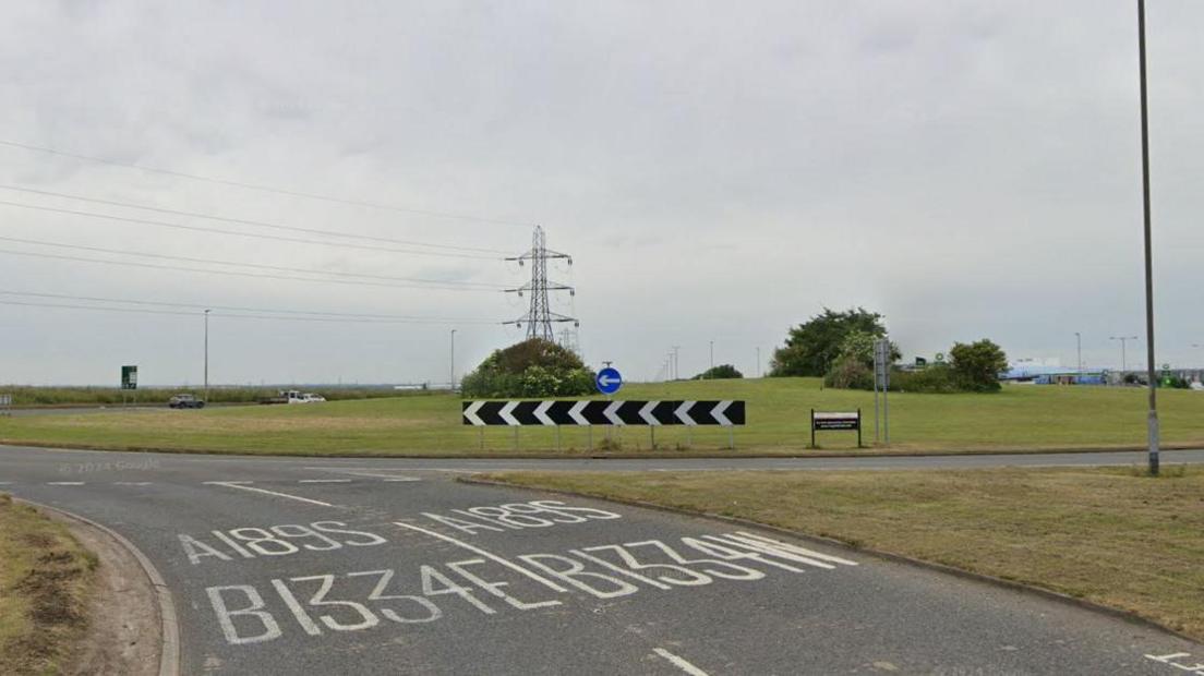 Streetview of a roundabout. There are two lanes approaching the roundabout, which is formed of a large green.