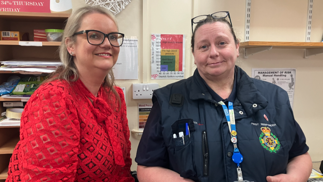 Two women look at the camera and are smiling. The woman on the left is wearing a red top, and has black rimmed glasses. The woman on the right has glasses perched on her head, and is wearing a blue top with the words first responder on it.