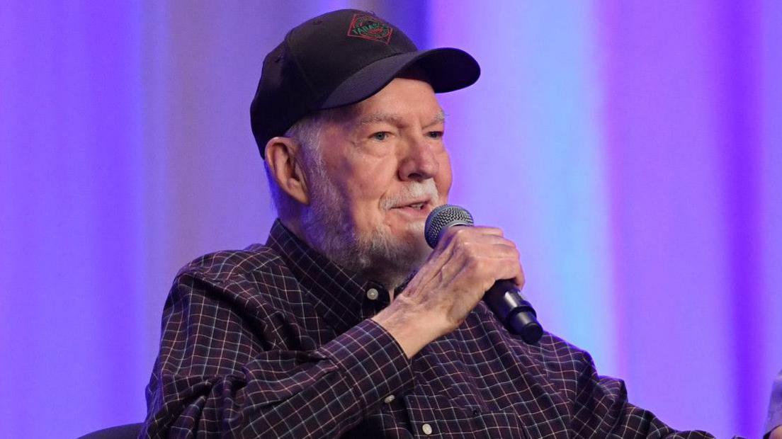 An image of Peter Renaday sitting in front of a purple curtain wearing a checked shirt and a cap, at an event in 2017