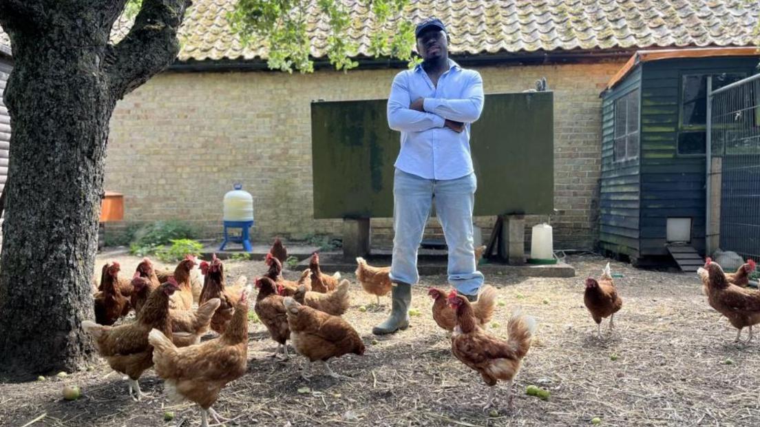 Fekky, wearing a navy blue cap, white shirt, jeans and wellington boots, stands with his arms folded looking at camera. He is surrounded by chickens on his farm.