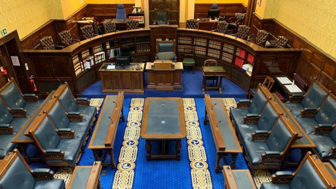 View looking down onto the empty Tynwald chamber, with both House of Keys and Legislative Council seats. The room has wooden benches, leather clad chairs, and a blue carpet with Celtic-style gold decoration.