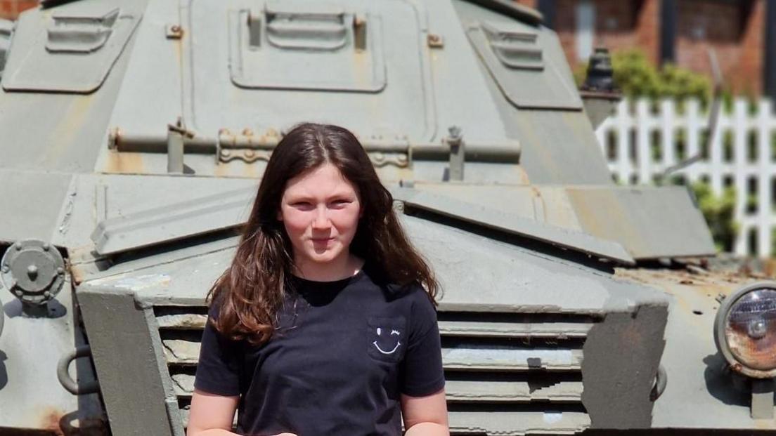 Maisie, in a black t-shirt and beige trousers, standing in front of a tank at the Heugh Battery Museum. She has brown hair parted in the middle. 