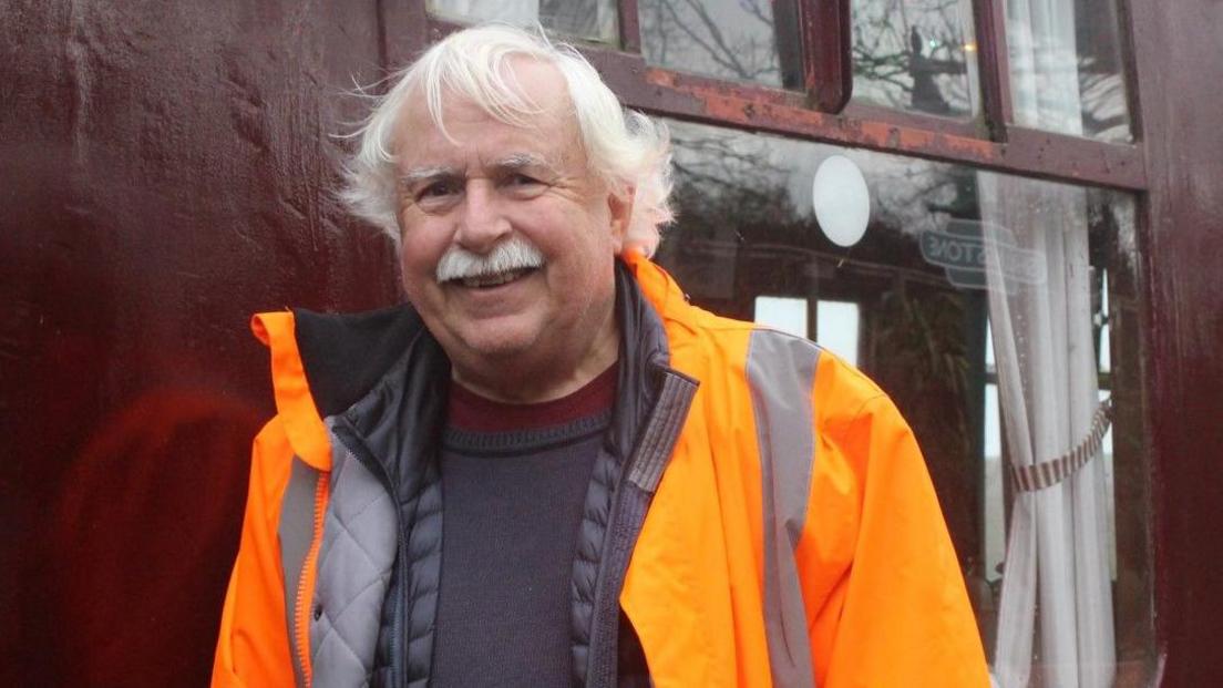 A smiling Phil Jacques wearing orange hi-viz overalls and standing in front of a dark red train carriage. He has white hair and a moustache.