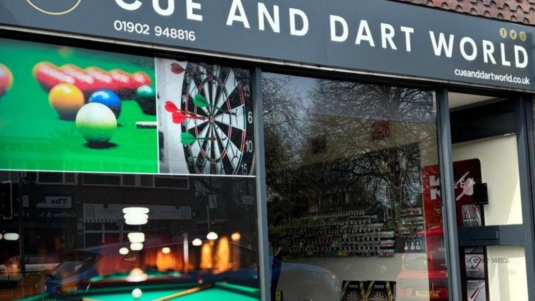 The outside of the shop, with large images of darts, snooker and pool. The name Cue and Dart World is at the top of the shop and the front door is open on the right.