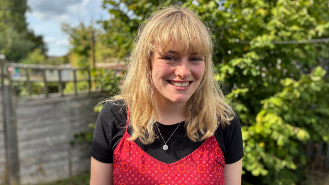 Rachael from the Wiltshire Centre for Independent Living smiling in a garden area. She is wearing a red top and black top and has blonde hair. There is a fence and a large green bush behind her. The sky is blue, with a grey cloud.
