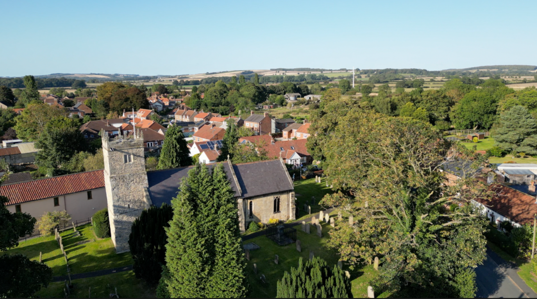 A bird's eye view of Shiptonthorpe, lush green, tall trees cast shadows over the village church. In the distance houses are nestled in between greenery.