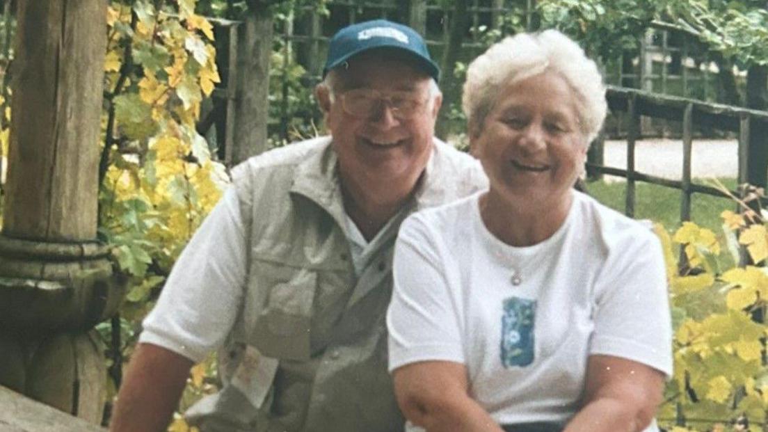 David and Pat Johnson looking to camera and smiling in a recent photo on holiday with a garden behind them 